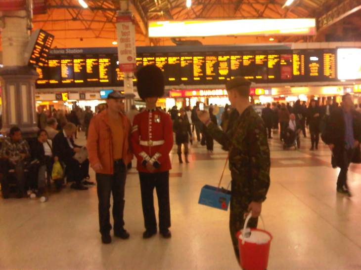 A soldier in uniform, carrying a poppy appeal bucket, takes a photo of a tourist standing next to another soldier (this one in red, gold and a bearskin hat), on the concourse of Victoria station.