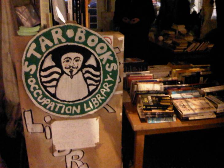 A table displaying books, and a large logo imitating starbucks but reading starbooks, with a Guy Fawkes mask in the middle.